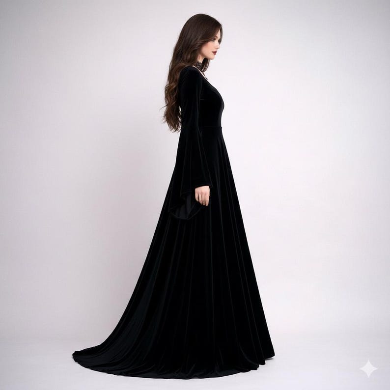 Pode incluir: Um vestido longo de veludo preto com uma saia fluida e mangas compridas e largas. O vestido tem um decote alto e uma cauda subtil. A modelo est&aacute; em perfil contra um fundo branco liso, mostrando a elegante silhueta do vestido.