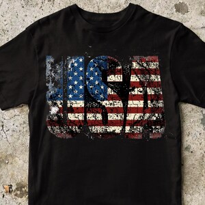 May include: Black t-shirt featuring a distressed design of the USA flag. The word "USA" is displayed in red, white, and blue, with a worn, vintage effect, suitable for casual wear.