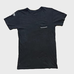 Puede incluir: Camiseta negra de manga corta con bolsillo en el pecho. El bolsillo tiene texto blanco. Hay texto blanco en la manga izquierda. La camiseta está hecha de un material suave.