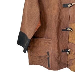 Może przedstawiać: Brązowa i beżowa kurtka z czarnym wykończeniem na rękawach. Kurtka ma kieszeń z motywem słońca i drewniane zapięcia na kołki. Tkanina wydaje się teksturowana.