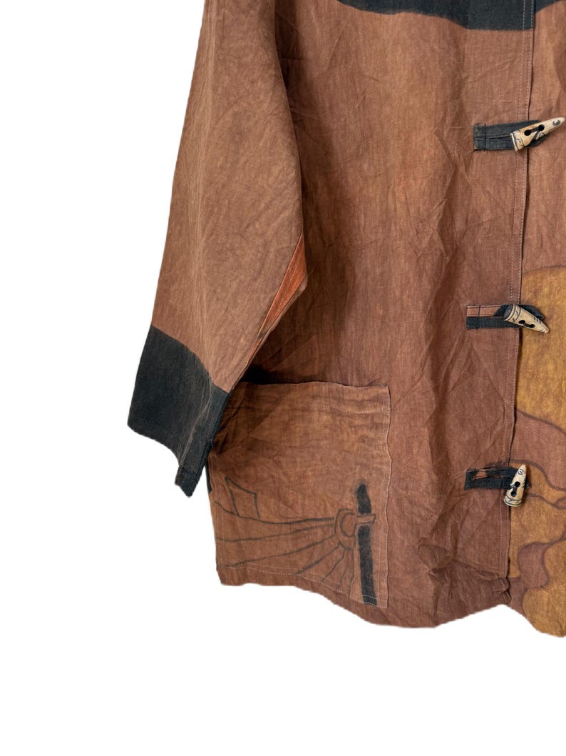 Może przedstawiać: Brązowa i beżowa kurtka z czarnym wykończeniem na rękawach. Kurtka ma kieszeń z motywem słońca i drewniane zapięcia na kołki. Tkanina wydaje się teksturowana.