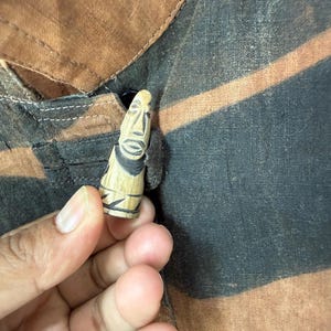 Może przedstawiać: Mała, rzeźbiona drewniana figurka ze stylizowaną twarzą, na tle brązowej i czarnej tkaniny. Figurka ma jasnobrązowy kolor z czarnymi detalami. Prawdopodobnie jest to przedmiot dekoracyjny lub unikalny guzik.