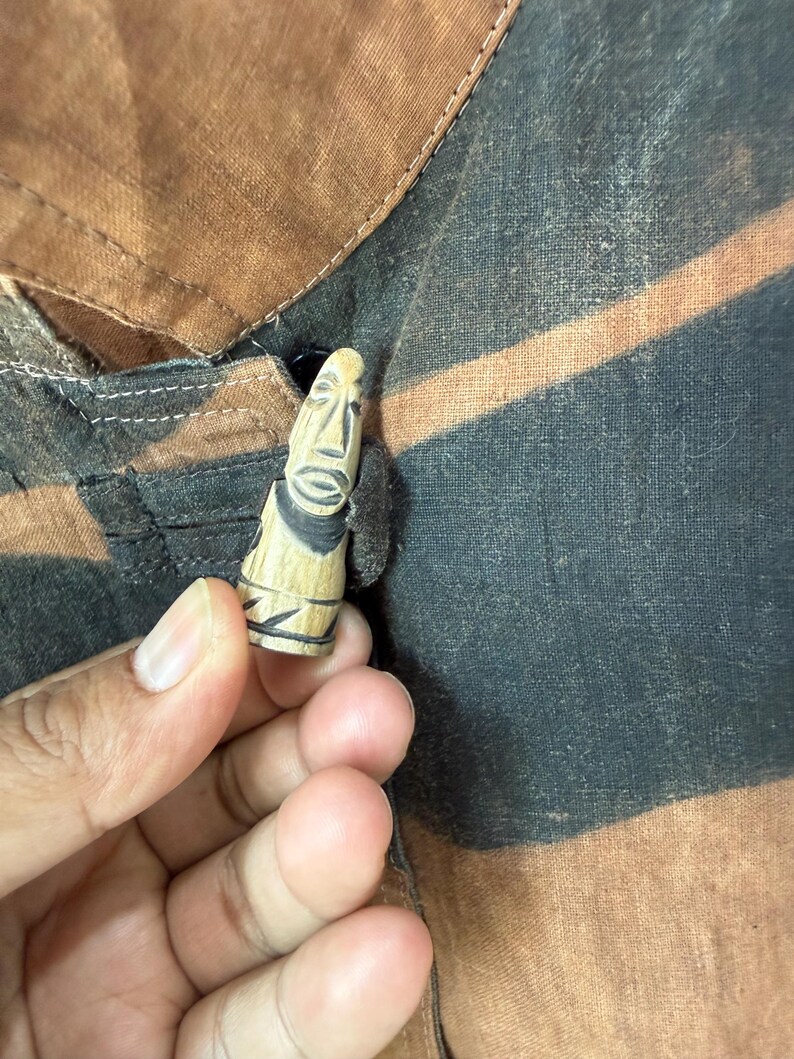 Może przedstawiać: Mała, rzeźbiona drewniana figurka ze stylizowaną twarzą, na tle brązowej i czarnej tkaniny. Figurka ma jasnobrązowy kolor z czarnymi detalami. Prawdopodobnie jest to przedmiot dekoracyjny lub unikalny guzik.