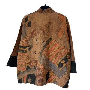 Może przedstawiać: Brązowa kurtka w stylu kimono ze szczeg&oacute;łową ilustracją samuraja na plecach. Grafika przedstawia odcienie brązu, pomarańczowego i czarnego. Rękawy mają czarne mankiety. Kurtka wisi na czarnym wieszaku.