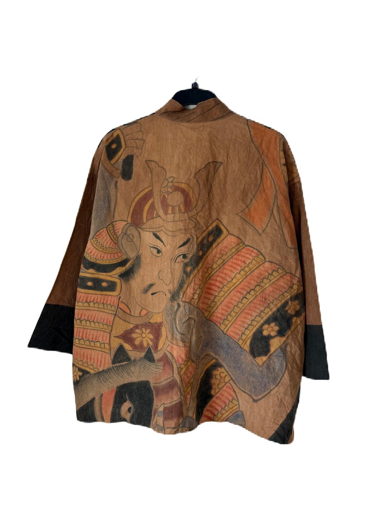 Może przedstawiać: Brązowa kurtka w stylu kimono ze szczeg&oacute;łową ilustracją samuraja na plecach. Grafika przedstawia odcienie brązu, pomarańczowego i czarnego. Rękawy mają czarne mankiety. Kurtka wisi na czarnym wieszaku.