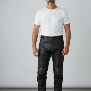Puede incluir: Pantalones de motorista de cuero negro con secciones de rodilla acolchadas. Los pantalones tienen una cintura alta y se combinan con una camiseta blanca y zapatillas blancas. El modelo está de pie frente a un fondo blanco liso.