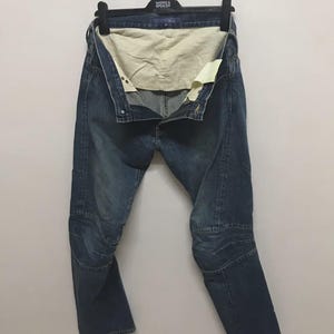 Puede incluir: Vaqueros de mezclilla azules con paneles de rodilla reforzados, colgados de una percha negra. Los vaqueros están desabrochados, revelando un forro interior de color claro. Los vaqueros tienen un diseño clásico de cinco bolsillos.