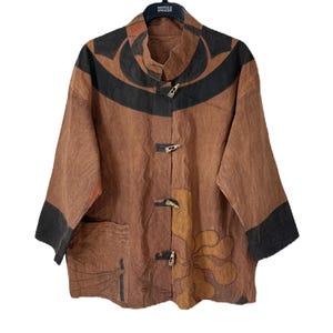 Może przedstawiać: Brązowa kurtka z czarnym i pomarańczowym abstrakcyjnym wzorem. Kurtka ma kołnierz mandaryński, trzy drewniane zapięcia na guziki i rękawy trzy czwarte z czarnymi mankietami. Kieszeń jest widoczna po lewej stronie.