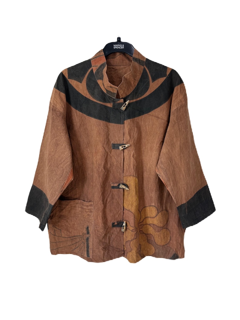 Może przedstawiać: Brązowa kurtka z czarnym i pomarańczowym abstrakcyjnym wzorem. Kurtka ma kołnierz mandaryński, trzy drewniane zapięcia na guziki i rękawy trzy czwarte z czarnymi mankietami. Kieszeń jest widoczna po lewej stronie.