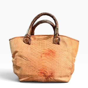 May include: A peach-colored handbag with a textured, scale-like pattern across the body. The bag features two dark brown, rounded handles attached with circular metal rings. The surface has subtle variations in tone and a visible, organic texture.