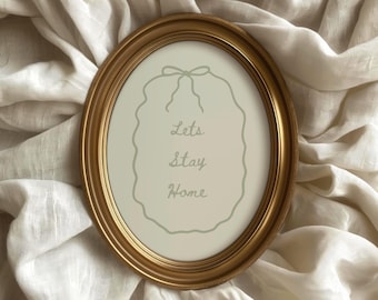 May include: An oval, gold-colored picture frame with the words "Lets Stay Home" in a soft green font. The frame is set against a backdrop of white fabric.