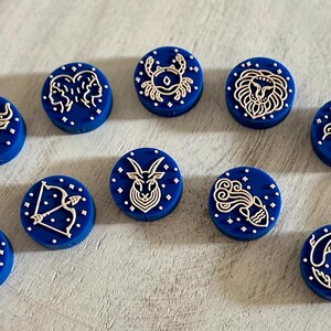 May include: A collection of twelve round, blue zodiac sign charms with white outlines. Each charm features a different zodiac symbol, such as a ram, a bull, and a crab. The charms are arranged on a light-colored surface.