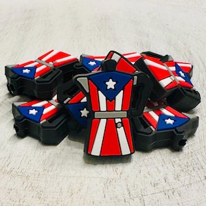 May include: A collection of black, red, white, and blue espresso machine-shaped charms. The charms feature the Puerto Rican flag design, with a white star on a blue background and red and white stripes. The charms are piled on a light-colored surface.