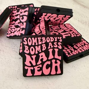 May include: A collection of square black charms featuring raised pink lettering that reads SOMEBODY'S BOMB ASS NAIL TECH. Each charm has a small circular hole at the top, designed for attachment to accessories or footwear with a flat, matte finish.