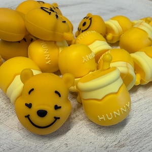 May include: A collection of yellow silicone beads shaped like Winnie the Pooh faces and honey pots with the word HUNNY embossed on the side. Each bead features a smooth matte finish, with black ink details for the eyes, nose, and smiling mouth.