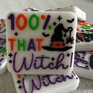 May include: White square soap bars with colorful Halloween-themed text and graphics. The bars feature the phrase "100% THAT Witch!" in orange, green, and purple. A witch hat and spider are also displayed.