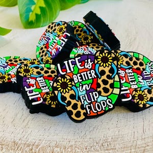 May include: A collection of colorful shoe charms with the phrase "LIFE is BETTER in FLIP FLOPS". The charms feature a leopard print design, sunflowers, and a variety of bright colors, perfect for decorating footwear.