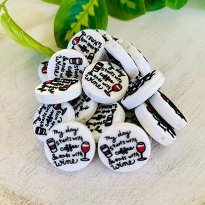 May include: A collection of white, round beads with black text and red accents. Each bead features the phrase "My day starts with coffee & ends with wine" along with coffee cup and wine glass illustrations. Some beads are cylindrical.