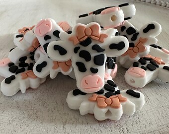 May include: A collection of white cow-shaped embellishments with black spots, pink noses, and peach-colored bows. These decorative pieces are designed with a flat back and are suitable for various craft projects.