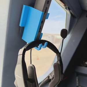 May include: A bright blue plastic mount holds a black electronic device vertically against a grey panel. Below the device, a matching blue plastic hook supports a pair of black Bose aviation headsets with a microphone boom and padded ear cups.