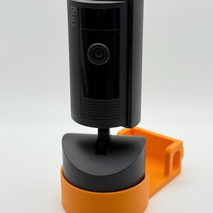 Op de afbeelding: Een zwarte beveiligingscamera met het woord "ring" aan de zijkant. De camera is gemonteerd op een zwarte basis met een oranje beugel. De camera heeft een lens en een luidspreker.