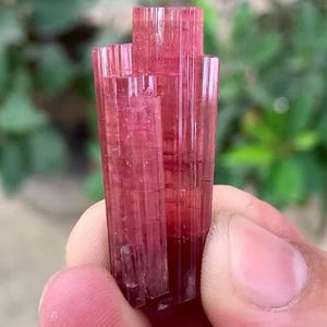 May include: A raw, elongated pink tourmaline crystal cluster featuring multiple vertical, striated columns with a translucent, deep rose-colored appearance. The mineral specimen displays natural geometric facets and a vibrant, consistent pink hue throughout its structure.