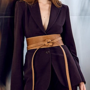 Puede incluir: Un traje de color ciruela oscuro con un cinturón ancho de cuero marrón atado a la cintura. El traje tiene una americana con cuello en V profundo y pantalones a juego. El cinturón añade un acento elegante.