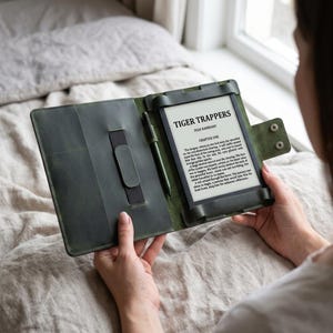 Puede incluir: Una funda de lector electrónico de cuero verde oscuro con un lápiz óptico a juego. El lector electrónico muestra el texto "TIGER TRAPPERS" de Ivan Basarriant. La funda tiene una correa para sujetar el lector y un bolsillo para el lápiz.