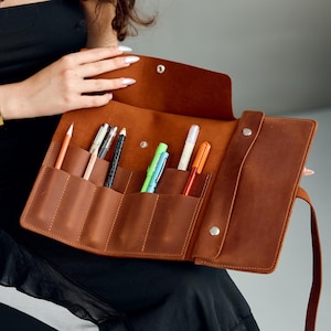 May include: Open, brown leather pen roll with multiple pockets holding various pens and pencils. The roll is secured with a strap and snap closure. The leather has a natural, textured appearance. The roll is partially open, revealing the writing instruments.