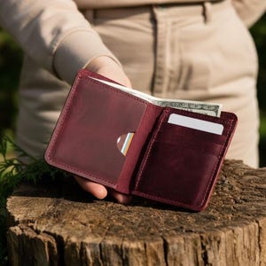 Può includere: Portafoglio in pelle bordeaux aperto con più fessure per carte e uno scomparto per contanti. Il portafoglio è tenuto aperto, rivelando carte e una banconota. Il portafoglio è su un ceppo d'albero.