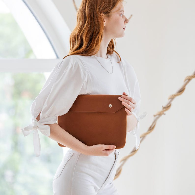 May include: A brown leather laptop sleeve with a flap closure and a silver press stud. The rectangular sleeve is made of smooth leather. The sleeve is being held in front of a person.