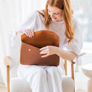 May include: A brown leather laptop bag with a flap closure and a silver snap. The bag is open, revealing the interior. The bag is held by a person wearing a white shirt and trousers. The bag appears to be approximately 38 cm wide.