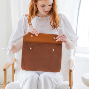 May include: A brown leather portfolio with a flap secured by two silver snap closures. The interior is a lighter shade of brown. The portfolio is held open, revealing the inside. The product is designed to carry documents or a laptop.