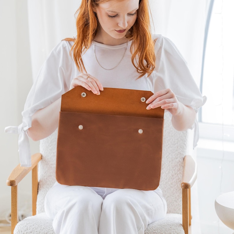 May include: A brown leather portfolio with a flap secured by two silver snap closures. The interior is a lighter shade of brown. The portfolio is held open, revealing the inside. The product is designed to carry documents or a laptop.