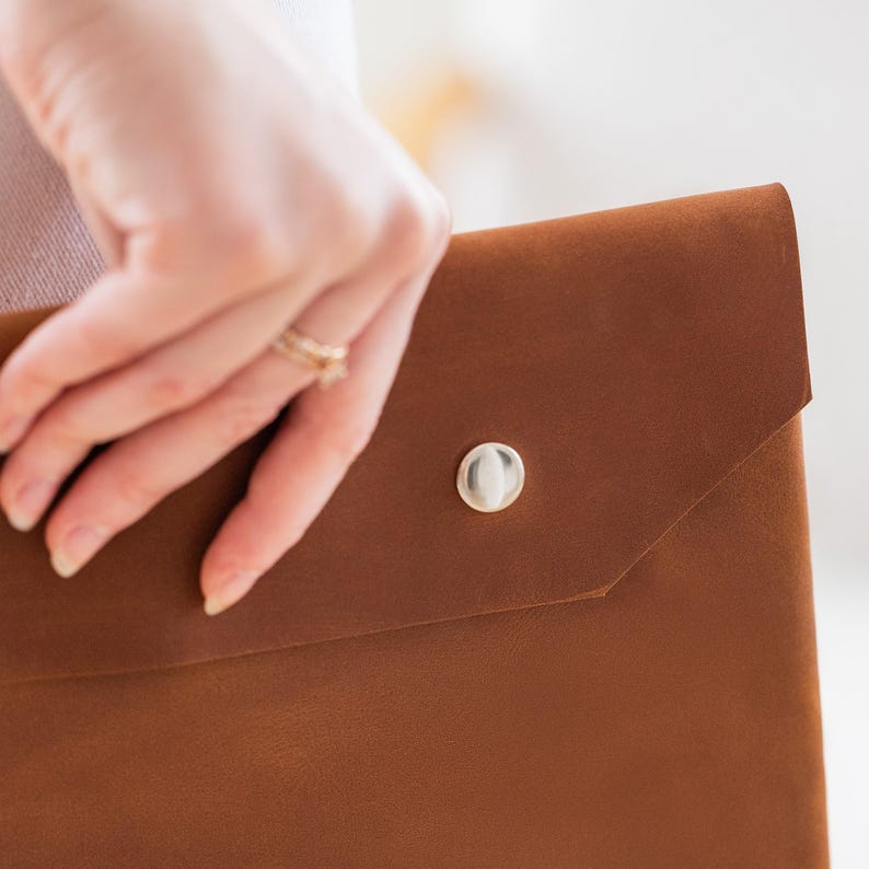 May include: A brown leather portfolio with a silver press stud closure. The portfolio features a minimalist design and is made of smooth, high-quality leather. The flap is secured with a round, silver-coloured press stud.