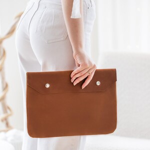 May include: A tan leather laptop sleeve with two silver press studs. The rectangular case is held by a person wearing white trousers. The sleeve has a simple, minimalist design, suitable for protecting a laptop or tablet.