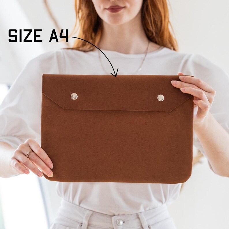May include: A brown A4-sized laptop sleeve with two silver snap closures. The text "SIZE A4" is visible in the upper left corner. The sleeve is made of a smooth material and has a flap closure.