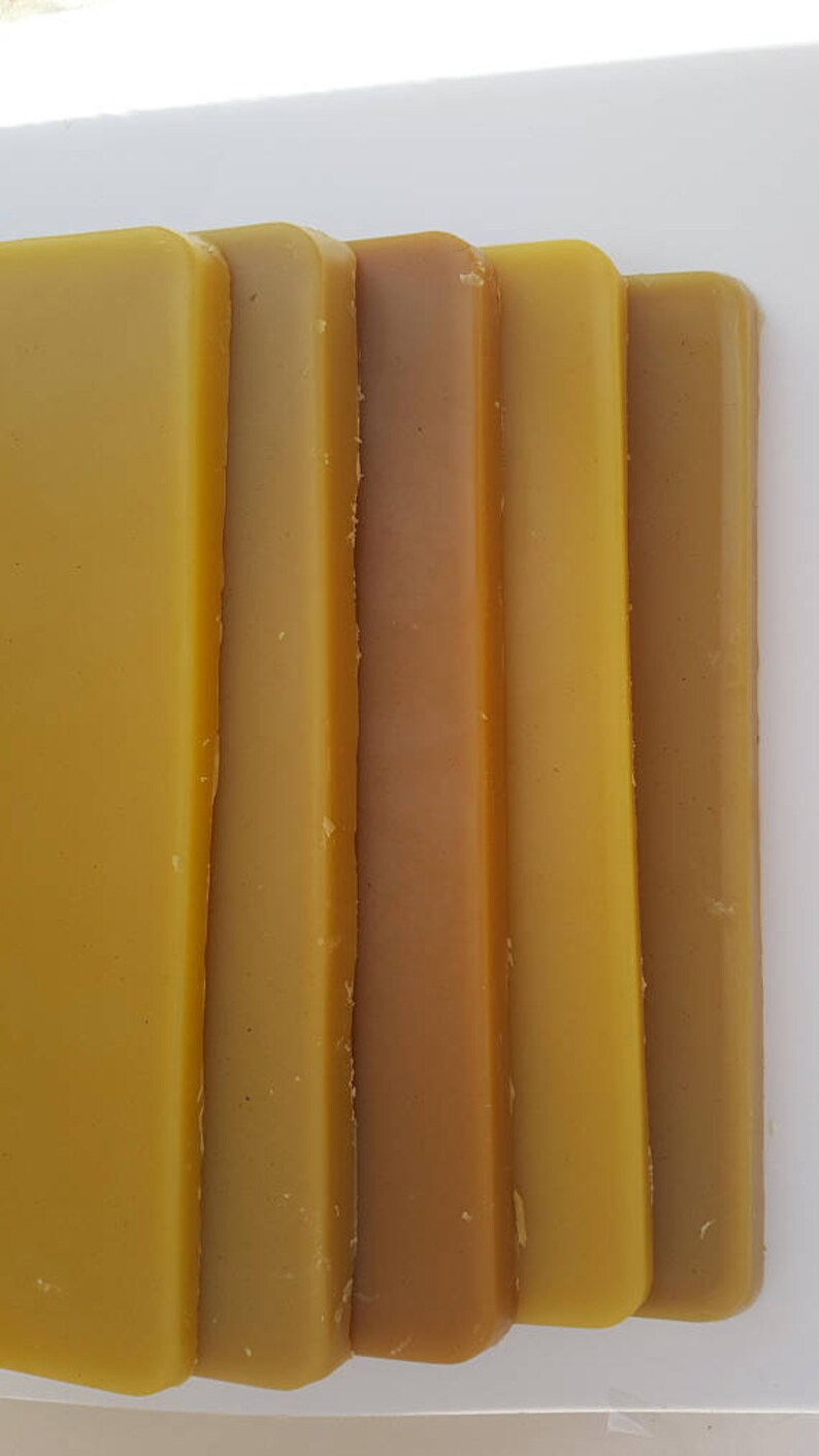 May include: Close-up of several rectangular bars of beeswax in varying shades of yellow and brown. The bars are arranged vertically, showcasing the natural color variations of the wax. The image highlights the texture and color of the beeswax.