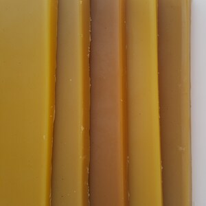 May include: Close-up of several rectangular bars of beeswax in varying shades of yellow and brown. The bars are arranged vertically, showcasing the natural color variations of the wax.