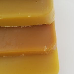 May include: Close-up of stacked, rectangular bars in varying shades of yellow and brown. The bars appear to be made of a solid, waxy material, with a smooth surface and slightly rounded edges. The colors range from a light, golden yellow to a deeper, amber hue.