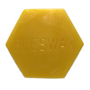 May include: A hexagonal block of yellow beeswax with the embossed word "BEESWAX". The wax has a smooth, slightly translucent appearance. The shape is a six-sided polygon, and the color is a warm, golden yellow.