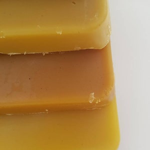 May include: Close-up of stacked, rectangular blocks of beeswax in varying shades of yellow and brown. The blocks have a smooth, slightly glossy surface, with a few small imperfections visible. The image highlights the natural color variations.