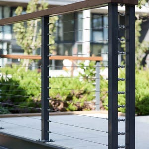 May include: Modern outdoor railing with dark gray metal posts and a brown wooden top rail. Horizontal stainless steel cables provide safety. The railing is set against a backdrop of green foliage and a building.