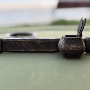 Peut inclure: Un ensemble de plumier antique avec des motifs floraux et géométriques complexes. L'ensemble comprend un petit pot d'encre à couvercle et un long repose-plume décoratif, le tout dans une finition métallique sombre et vieillie. L'encrier est ouvert.