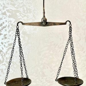May include: An antique brass balance scale with two oval pans hanging by chains from a horizontal beam. The scale is suspended from a central hook. The background is a textured, off-white surface.