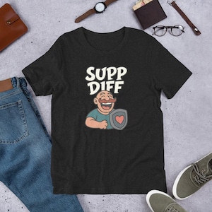 Puede incluir: Camiseta gris oscuro con las palabras blancas "SUPP DIFF" sobre una ilustración de dibujos animados de un hombre con bigote sosteniendo un escudo con un corazón. La camiseta es de material suave.
