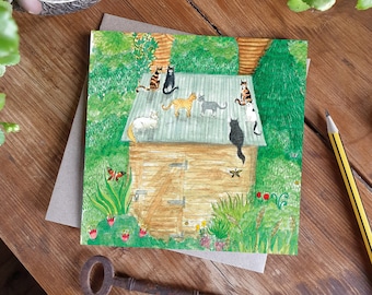 May include: A square greetings card with a watercolour illustration of a wooden shed, with cats on the roof and in the garden. The cats are various colours, including ginger, black, and white. The card is on a brown envelope.