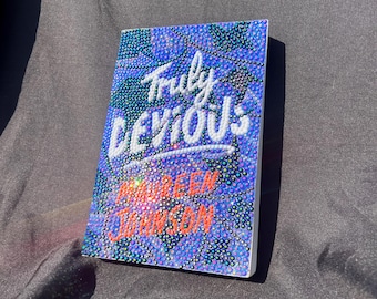 May include: A hardcover book titled "Truly Devious" by Maureen Johnson. The cover is a mosaic of blue, purple, and iridescent rhinestones. The title is in white script, and the author's name is in orange.
