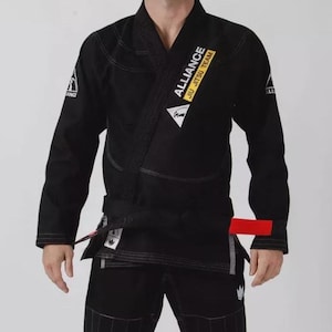 Puede incluir: Un kimono de jiu-jitsu negro con solapa gruesa y mangas largas. El pecho muestra el texto blanco ALLIANCE JIU JITSU TEAM sobre un fondo rectangular amarillo. Un cinturón negro con una lengüeta roja está atado a la cintura. Costuras visibles.