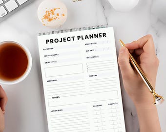 May include: A white spiral-bound project planner with the title "PROJECT PLANNER" in bold black letters. The planner includes sections for category, project name, objectives, resources, notes, and an action plan. A gold pen rests on the page.